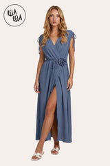 Front view of a model wearing the Denim Blue Flower Corsage Wrap Maxi Dress, showcasing the muted indigo-hued drape and the 3D floral waist accent.