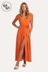 Front view of a model wearing the Orange Flower Corsage Wrap Maxi Dress, showcasing the vibrant citrus-hued drape and the 3D floral waist accent.