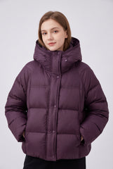 Front model view of a plum cropped hooded puffer jacket with a raised drawstring neckline, snap-button placket and side pockets, styled with black trousers.