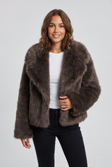 Front model view of a chocolate brown short faux fur jacket with an oversized fold-over collar and long sleeves, styled with a white top and black trousers.