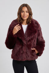 Front model view of a burgundy short faux fur jacket with an oversized fold-over collar, long sleeves and pockets, styled with black trousers.