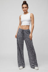 Full-body model view of grey sparkling wide-leg sequin trousers with a drawstring waistband, styled with a white sleeveless crop top and white trainers.