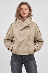 Front model view of a beige cropped double-breasted trench jacket with a large collar, shoulder tabs, buckled cuffs and side pockets.