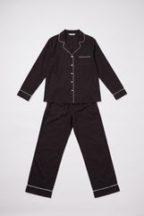 Flat product view of a two-piece black pyjama set showing the long sleeve button-through shirt, white piping, chest pocket and full-length matching trousers.