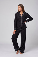Model wearing a black long sleeve pyjama set with white contrast piping, a notched collar, button-through shirt, chest pocket and matching long trousers.
