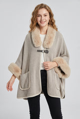 Front model view of the beige textured knit poncho cape with faux fur collar and cuffs, buckle fastening and two patch pockets.